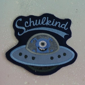 Könnte beinhalten: Blau-grauer gestickter Aufnäher mit einem Comic-Alien in einem Raumschiff. Der Aufnäher trägt die Aufschrift "Schulkind".