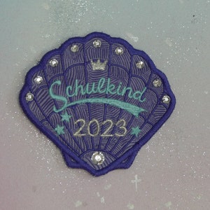 Op de afbeelding: Paarse geborduurde schelp patch met strass steentjes, een kroon en de tekst "Schulkind 2023".