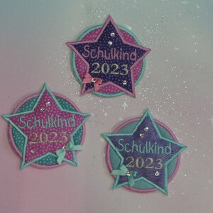 Könnte beinhalten: Drei sternförmige Patches in Lila, Rosa und Türkis mit dem in Gold gestickten Text "Schulkind 2023". Jedes Patch hat eine kleine Schleife und Strasssteine.