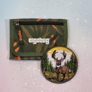 Könnte beinhalten: Ein rechteckiges olivgrünes Portemonnaie mit orangefarbenen Batik-Akzenten und dem weißen Schriftzug "ergobag" auf der Vorderseite, dazu ein runder gestickter Aufnäher mit einem braunen Hirsch in einem Wald aus grünen Tannen vor gelbem Himmel.