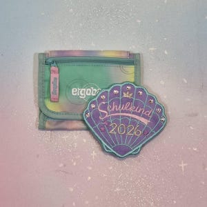 Peut inclure: Un portefeuille tie-dye avec une fermeture éclair et la marque "ergob" est à côté d'un écusson en forme de coquillage violet. L'écusson porte le texte "Schulkind 2026" et est orné de strass.