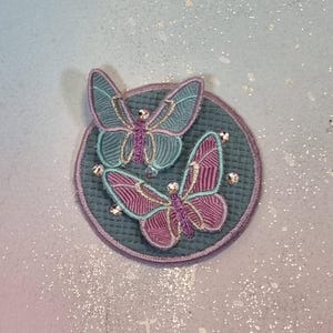 Könnte beinhalten: Gestickter Aufnäher mit zwei Schmetterlingen in Türkis und Rosa, mit lila Details. Die Schmetterlinge sind mit kleinen, funkelnden Strasssteinen verziert und befinden sich auf einem türkisfarbenen, kreisförmigen Hintergrund mit lila Rand.