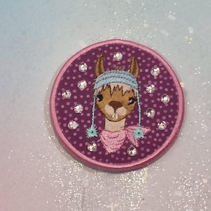 May include: Embroidered patch featuring a llama wearing a blue and white knit hat and a pink scarf. The llama is centered on a purple polka-dotted background, surrounded by clear rhinestones, and framed by a pink border. This patch is suitable for clothing or accessories.