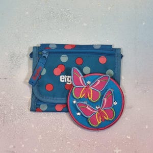 Op de afbeelding: Een teal portemonnee met een ritssluiting en een flap, versierd met rode en roze stippen. Een cirkelvormige patch met twee roze vlinders en strass-accenten ligt erop. De portemonnee heeft het woord "erg" erop gedrukt.