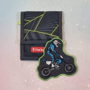 Op de afbeelding: Een donkergrijze portemonnee met een geometrisch ontwerp en een neon groen accent. Een patch van een motorcrosser op een motorfiets staat naast de portemonnee. De portemonnee heeft de tekst "Step by".