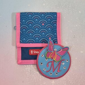 Könnte beinhalten: Ein blaues Portemonnaie mit rosa Einfassung und einem passenden Schmetterlingsaufnäher. Das Portemonnaie hat ein sich wiederholendes Muster aus weißen Punkten und Halbkreisen. Der Aufnäher ist hellblau mit einem rosa Schmetterling und dem Buchstaben "M".