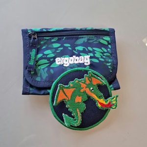Könnte beinhalten: Ein marineblaues Portemonnaie mit grünem Blättermuster und dem Markennamen "ergobag" in Weiß. Ein kreisförmiger Aufnäher mit einem grünen und orangefarbenen Drachendesign befindet sich neben dem Portemonnaie. Der Drache speit Feuer.