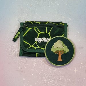 Könnte beinhalten: Eine grüne Geldbörse und ein runder Aufnäher. Die Geldbörse hat ein geometrisches Muster und den Markennamen "ergobag". Der Aufnäher zeigt ein Baumdesign mit braunem Stamm und grünen Blättern.