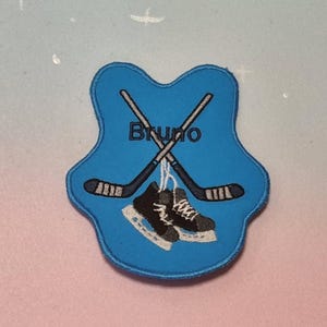 Puede incluir: Parche de fieltro azul con un par de patines de hockey y dos palos de hockey cruzados. El nombre "Bruno" está bordado en blanco en el parche.