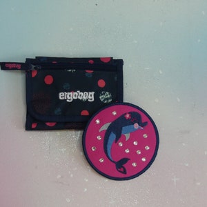 May include: A dark blue wallet with red polka dots and the white text ergobag, paired with a round, bright pink patch featuring a blue dolphin graphic, a small flower detail, and several clear, reflective circular embellishments.
