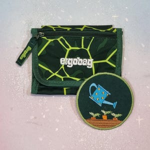 Könnte beinhalten: Eine grün-gelb gemusterte Geldbörse mit dem Markennamen "ergobag" in Weiß. Ein passender runder Aufnäher zeigt ein Gießkannen-Design über Karotten. Die Geldbörse hat einen Reißverschluss und ein kleines Etikett mit dem Markennamen.