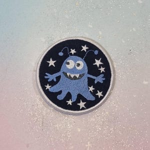 Könnte beinhalten: Gestickter Aufnäher mit einem hellblauen Cartoon-Monster mit breitem Grinsen, zwei Antennen und kleinen Armen. Das Monster ist von weißen Sternen auf einem marineblauen kreisförmigen Hintergrund umgeben. Der Aufnäher hat einen weißen Rand.