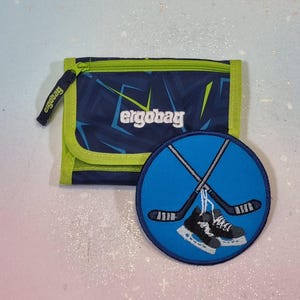 Könnte beinhalten: Eine blau-grüne Geldbörse mit dem Aufdruck "ergobag" daneben ein blaues rundes Patch mit gekreuzten Hockeyschlägern und Schlittschuhen. Die Geldbörse hat einen Reißverschluss.