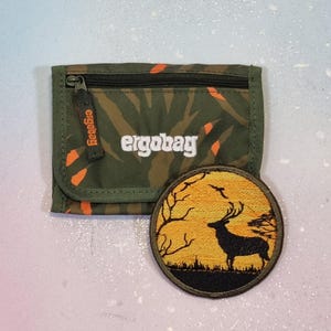 Op de afbeelding: Een rechthoekige olijfgroene portemonnee met een oranje bladerpatroon en de witte tekst ergobag, voorzien van een ritssluiting. Daarnaast ligt een ronde geborduurde patch met het zwarte silhouet van een hert tegen een gele zonsondergang.