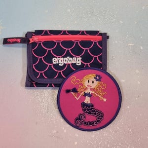 Könnte beinhalten: Ein marineblaues und pinkfarbenes Portemonnaie mit Reißverschluss und dem Aufdruck "ergobag", neben einem runden pinkfarbenen Aufnäher mit einem Meerjungfrau-Design. Das Portemonnaie hat ein Schuppenmuster.