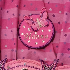 Op de afbeelding: Roze stof met een herhalend sterrenpatroon. Een cirkelvormige patch met de naam "Hanna" geborduurd in zilveren draad is versierd met een vlinderapplicatie en strass-steentjes. De patch is omrand met paars.