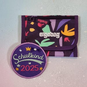 Peut inclure: Un portefeuille violet avec un motif abstrait coloré et le nom de la marque "ergobag" en blanc. À côté, un écusson rond avec le texte "Schulkind 2025" en écriture cursive, avec une couronne et des étoiles, le tout en violet, vert et or.