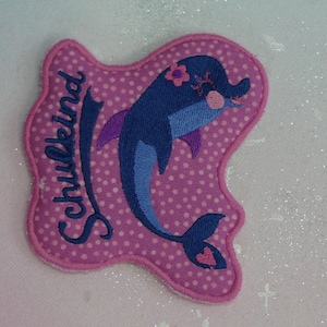 Könnte beinhalten: Rosa Stoff-Patch mit einem blauen und violetten Delfin mit einer Blume auf dem Kopf. Der Patch sagt "Schulkind" in blauen Buchstaben.