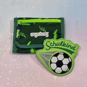 Könnte beinhalten: Grüne und schwarze Geldbörse mit dem Wort "ergobag" in Weiß. Die Geldbörse hat einen Reißverschluss und eine grüne Umrandung. Ein grüner Aufnäher mit einem Fußball und dem Wort "Schulkind" liegt neben der Geldbörse.