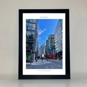 May include: A framed photograph of a city street in London, England. The street is lined with tall buildings and there is a red double-decker bus driving down the street. The text "Bishopsgate" and the coordinates "51°31' 13.9"N, 0° 4' 43.6"W" are printed at the top and bottom of the image.