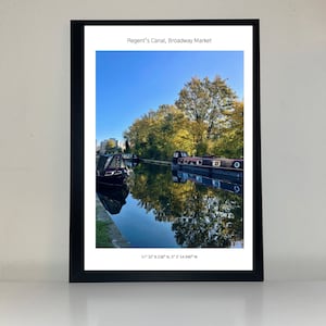 May include: A photograph of Regent's Canal in London, England. The canal is lined with trees and there are two boats docked on the water. The water is calm and reflects the sky and trees. The text "Regent's Canal, Broadway Market" and the coordinates "51° 32' 8.538" N, 0° 3' 54.940" W" are printed below the image.