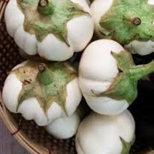 May include: A group of white eggplants with green tops in a woven basket. The eggplants are round and have a smooth, white skin.