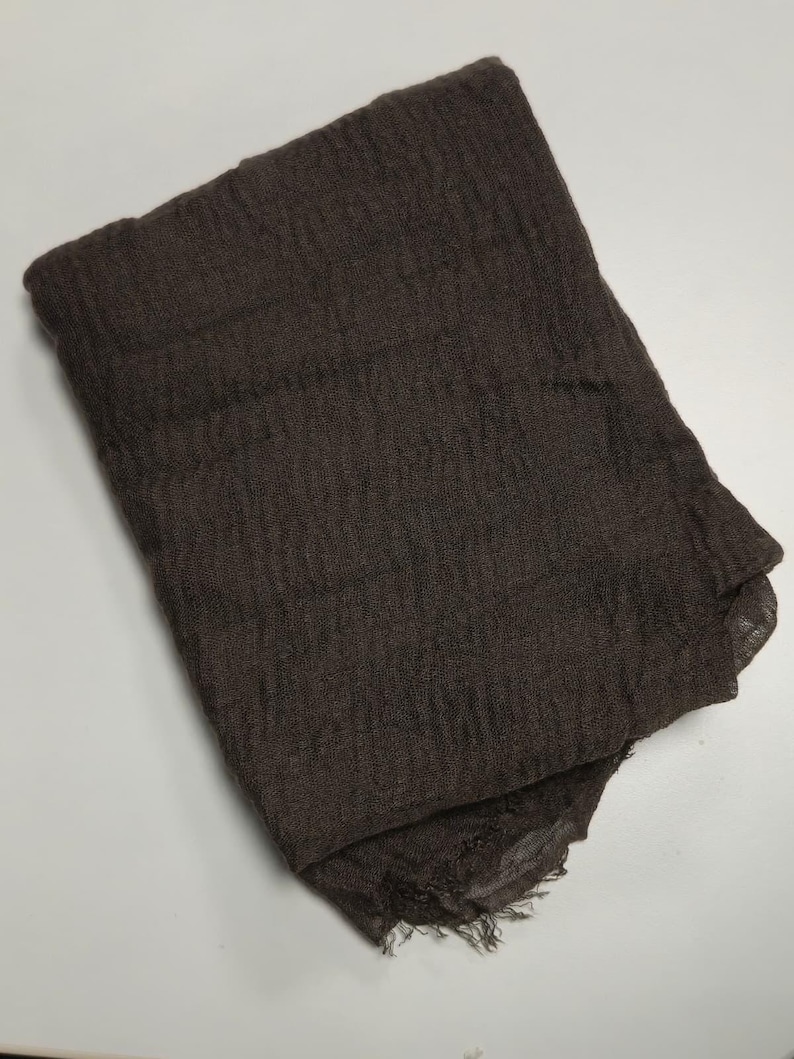 K&ouml;nnte beinhalten: Ein dunkelbrauner, rechteckiger Schal mit einer geknitterten, strukturierten Stoffoberfl&auml;che. Der Schal ist kompakt gefaltet und weist am unteren Saum ausgefranste, unvers&auml;uberte Kanten auf, die ein leichtes und halbtransparentes Gewebe zeigen.