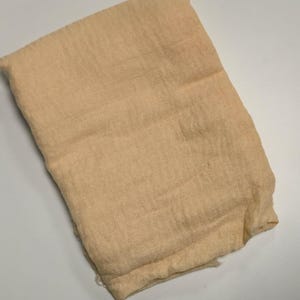 K&ouml;nnte beinhalten: Ein rechteckiges St&uuml;ck hellbeiger Stoff mit einer sichtbaren Webstruktur. Das Material wirkt weich und leicht und weist eine leicht zerknitterte Oberfl&auml;che sowie gefaltete Kanten auf. Das Textil ist in einer einfachen, flachen Form gefaltet.