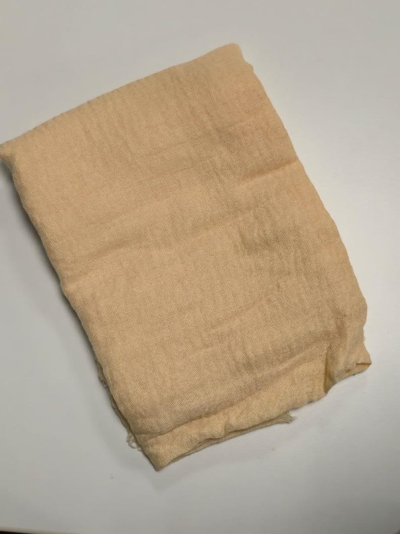 K&ouml;nnte beinhalten: Ein rechteckiges St&uuml;ck hellbeiger Stoff mit einer sichtbaren Webstruktur. Das Material wirkt weich und leicht und weist eine leicht zerknitterte Oberfl&auml;che sowie gefaltete Kanten auf. Das Textil ist in einer einfachen, flachen Form gefaltet.