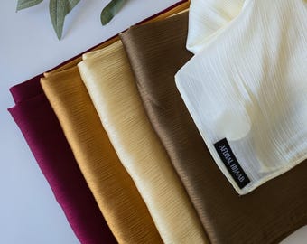 May include: A stack of five rectangular hijabs in shades of burgundy, mustard yellow, light gold, bronze, and cream. Each hijab features a fine, vertical crinkle texture. The cream-colored hijab has a black fabric label reading AFDHAL HIJAAB.