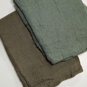 K&ouml;nnte beinhalten: Zwei gefaltete rechteckige Stoffst&uuml;cke mit einer sichtbaren, knittrigen Textur. Ein St&uuml;ck ist in einem ged&auml;mpften Salbeigr&uuml;n gehalten, w&auml;hrend das andere einen dunkleren olivbraunen Farbton aufweist. Beide Artikel wirken weich und leicht.