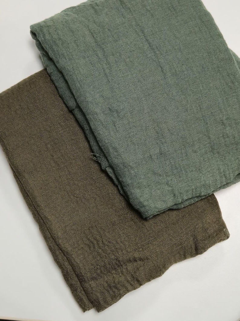 K&ouml;nnte beinhalten: Zwei gefaltete rechteckige Stoffst&uuml;cke mit einer sichtbaren, knittrigen Textur. Ein St&uuml;ck ist in einem ged&auml;mpften Salbeigr&uuml;n gehalten, w&auml;hrend das andere einen dunkleren olivbraunen Farbton aufweist. Beide Artikel wirken weich und leicht.