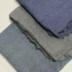K&ouml;nnte beinhalten: Drei rechteckige Schals mit einer geknitterten, strukturierten Stoffoberfl&auml;che. Der oberste Schal ist in tiefem Marineblau, der mittlere in neutralem Anthrazitgrau und der unterste in ged&auml;mpftem Schieferblau gehalten. Alle haben ausgefranste Kanten.