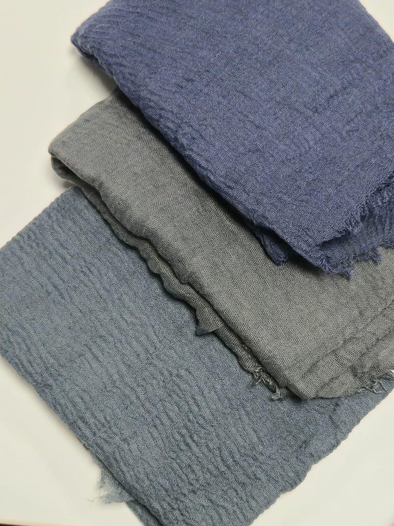 K&ouml;nnte beinhalten: Drei rechteckige Schals mit einer geknitterten, strukturierten Stoffoberfl&auml;che. Der oberste Schal ist in tiefem Marineblau, der mittlere in neutralem Anthrazitgrau und der unterste in ged&auml;mpftem Schieferblau gehalten. Alle haben ausgefranste Kanten.