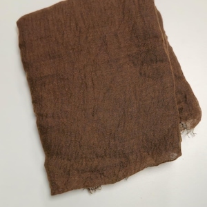K&ouml;nnte beinhalten: Ein rechteckiger brauner Schal mit einer leichten, halbtransparenten und knittrigen Textur. Der Stoff weist eine lockere Webart mit sichtbaren horizontalen und vertikalen F&auml;den auf, und die Kanten haben ein leicht ausgefranstes Aussehen.