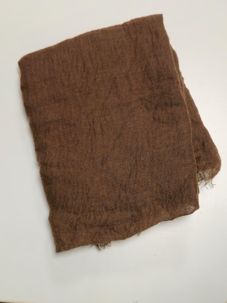 K&ouml;nnte beinhalten: Ein rechteckiger brauner Schal mit einer leichten, halbtransparenten und knittrigen Textur. Der Stoff weist eine lockere Webart mit sichtbaren horizontalen und vertikalen F&auml;den auf, und die Kanten haben ein leicht ausgefranstes Aussehen.