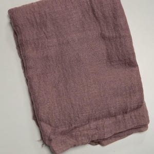 K&ouml;nnte beinhalten: Ein gefaltetes rechteckiges Stoffst&uuml;ck in einem ged&auml;mpften, staubigen Mauveton. Das Textil weist eine sichtbare, locker gewebte Gaze-Struktur mit einem leicht zerknitterten Erscheinungsbild auf der gesamten Oberfl&auml;che auf und wirkt einheitlich.