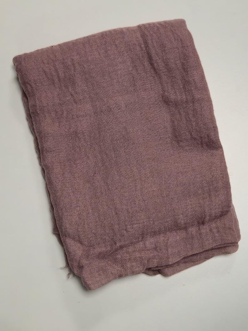 K&ouml;nnte beinhalten: Ein gefaltetes rechteckiges Stoffst&uuml;ck in einem ged&auml;mpften, staubigen Mauveton. Das Textil weist eine sichtbare, locker gewebte Gaze-Struktur mit einem leicht zerknitterten Erscheinungsbild auf der gesamten Oberfl&auml;che auf und wirkt einheitlich.
