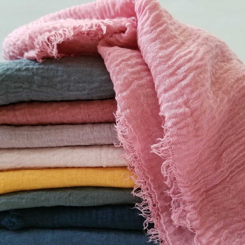 K&ouml;nnte beinhalten: Ein Stapel gefalteter rechteckiger Stoffschals mit einer geknitterten, strukturierten Oberfl&auml;che und ausgefransten Kanten. Die Kollektion umfasst verschiedene Farben wie Altrosa, Dunkelgrau, Mauve, Hellrosa, Senfgelb, Petrol und tiefes Marineblau.