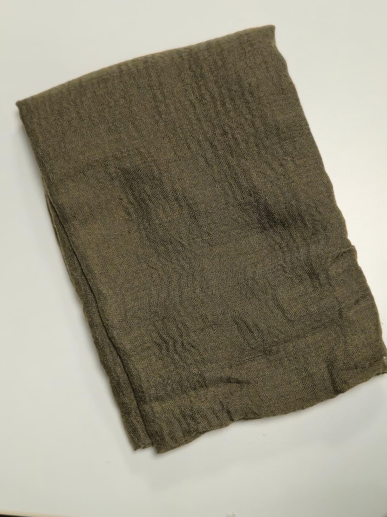 K&ouml;nnte beinhalten: Ein rechteckiges St&uuml;ck Stoff in einem einfarbigen Olivgr&uuml;n. Das Textil weist eine ausgepr&auml;gte, zerknitterte und strukturierte Oberfl&auml;che mit sichtbaren Falten und einer lockeren Weboptik auf. Die Kanten sind gerade und sauber verarbeitet.