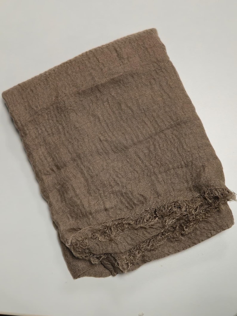 K&ouml;nnte beinhalten: Ein rechteckiger Schal in einem einfarbigen Taupe-Braun. Der Stoff weist durchgehend eine ausgepr&auml;gte, zerknitterte Textur auf. Die untere Kante des Schals ist mit einem ausgefransten Saum versehen, der dem Design ein haptisches Element verleiht.