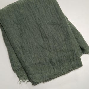 K&ouml;nnte beinhalten: Ein gefalteter rechteckiger Schal in ged&auml;mpftem Olivgr&uuml;n. Der Stoff weist eine ausgepr&auml;gte, knittrige Textur mit lockerer Webart und ausgefransten Kanten am unteren Saum auf, wobei das gesamte St&uuml;ck einen einheitlichen, monochromen Farbton zeigt.