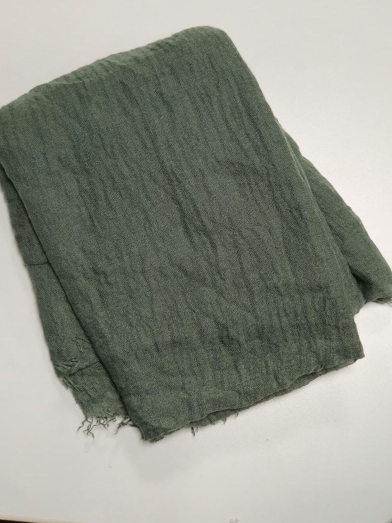K&ouml;nnte beinhalten: Ein gefalteter rechteckiger Schal in ged&auml;mpftem Olivgr&uuml;n. Der Stoff weist eine ausgepr&auml;gte, knittrige Textur mit lockerer Webart und ausgefransten Kanten am unteren Saum auf, wobei das gesamte St&uuml;ck einen einheitlichen, monochromen Farbton zeigt.