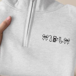 Puede incluir: Una sudadera gris claro con cremallera de cuarto de cremallera y cremallera plateada. La sudadera presenta un pequeño diseño bordado de lazos y otras formas en hilo negro. La prenda está doblada y sostenida por una mano.