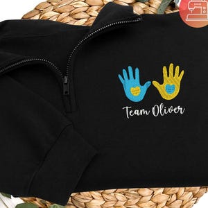 May include: A black quarter-zip sweatshirt featuring an embroidered design of two handprints, one light blue and one yellow, each containing a small heart. Below the handprints, the text Team Oliver is embroidered in white cursive script.