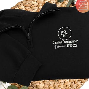 Puede incluir: Sudadera negra con cremallera de cuarto de cremallera con el texto "Cardiac Sonographer Jessica, RDCS" en blanco. El diseño incluye un gráfico circular blanco con una ilustración de corazón. La sudadera está doblada sobre una cesta tejida.