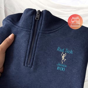 May include: Navy blue quarter-zip sweatshirt with the text "Rad Tech" and a skeleton graphic embroidered in light blue. The name "Jessica RT(R)" is also embroidered below the graphic. The sweatshirt has a zipper and a high collar.