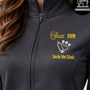 Może przedstawiać: Szara kurtka zapinana na zamek z haftowanymi detalami. Kurtka ma napis "Suzi DVM" w żółtym kolorze, grafikę strzykawki i śladu łapy oraz napis "Smile Vet Clinic" w kolorze żółtym. W prawym górnym rogu znajduje się haftowane logo.