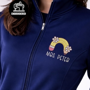 Puede incluir: Chaqueta azul marino con cremallera y diseño bordado de lápiz y flores. La chaqueta presenta el texto "MRS. PETER" bordado debajo del diseño. La palabra "EMBROIDERED" está en un círculo en la parte superior izquierda. La palabra "navy" está en la parte inferior izquierda.
