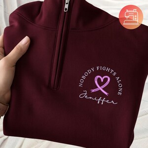 May include: A maroon quarter-zip sweatshirt featuring a light purple awareness ribbon shaped like a heart. The text reads NOBODY FIGHTS ALONE in a circular arc above the ribbon, with the name Jeniffer written in white cursive script below it.